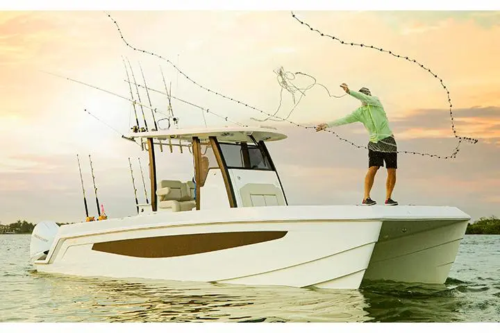 A man in a green shirt throws a fishing net from a white boat, surrounded by water at sunset, with fishing rods visible.